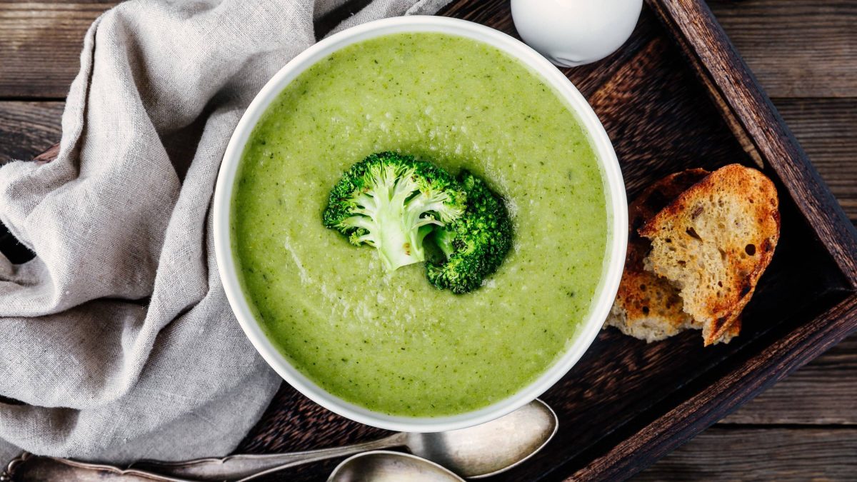 Pürierte Brokkoli-Zitronen-Suppe mit Kokosmilch und Ingwer in einer Schale, garniert mit frischem ganzen Brokkolieröschen und dazu frisch gebackenes Brot auf hölzernem Untergrund