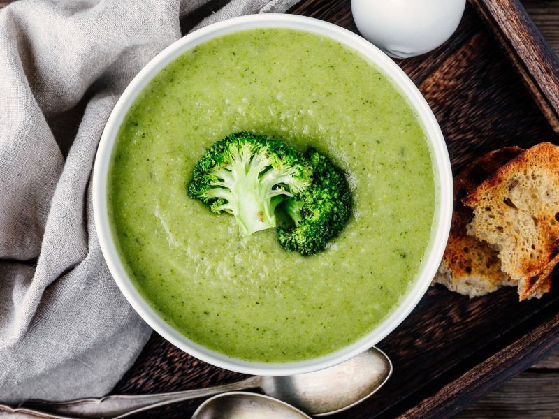 Pürierte Brokkoli-Zitronen-Suppe mit Kokosmilch und Ingwer in einer Schale, garniert mit frischem ganzen Brokkolieröschen und dazu frisch gebackenes Brot auf hölzernem Untergrund