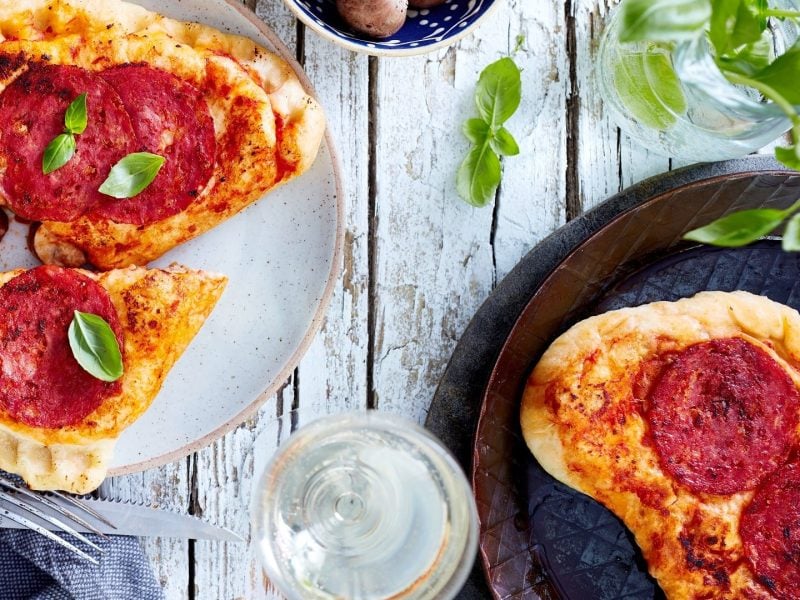 Calzone-Pfannenpizza auf einem Teller und in der Pfanne. Daneben Gläser mit verschiedenen Zutaten. Das Ganze auf einem hellen hölzernen Untergrund.