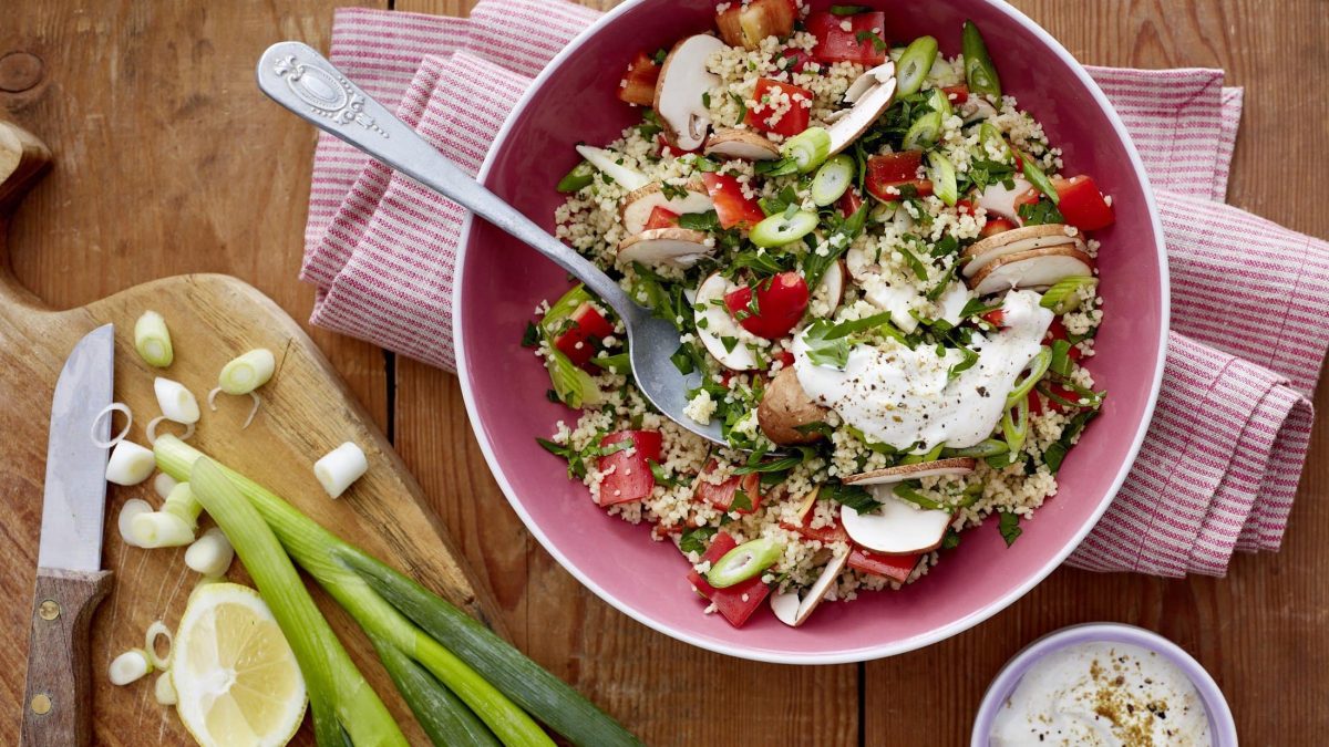 Couscous-Salat mit Pilzen in einer rosa Schüssel. Daneben verschiedenes Besteck und verschiedene Zutaten. Alles auf einem hölzernen Untergrund mit einem Küchentuch.