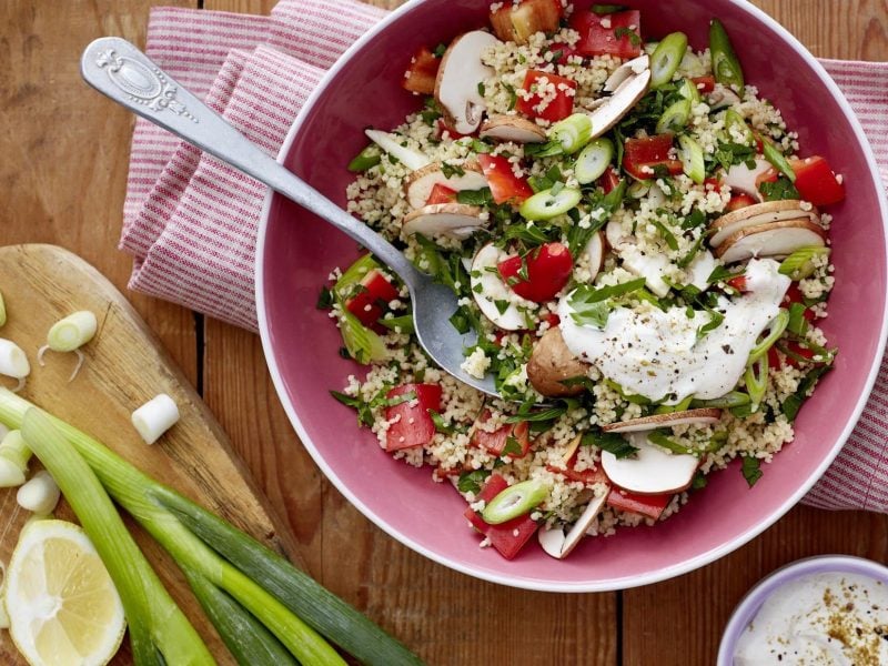 Couscous-Salat mit Pilzen in einer rosa Schüssel. Daneben verschiedenes Besteck und verschiedene Zutaten. Alles auf einem hölzernen Untergrund mit einem Küchentuch.