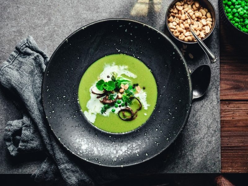 Ein schwarzer Teller Erbsen-Kokos-Suppe mit Minze, Erdnüssen und karamellisierten Zwiebelringen in der Draufsicht. Daneben stehen Schüsseln mit Zutaten.