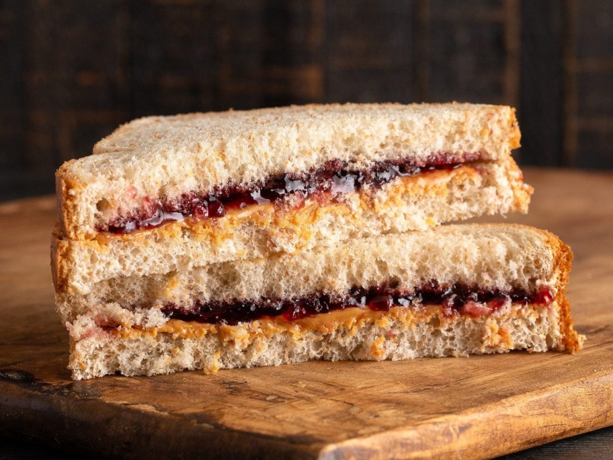 Ein Erdnussbutter-Marmeladen-Sandwich liegt halbiert und gestapelt auf einem Holzbrett.