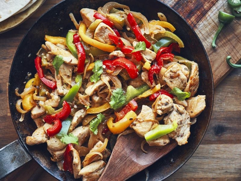 Fajita de Pollo in einer gusseisernen Pfanne. Daneben Chili und Guacamole. Das Ganze auf einem hölzernen Untergrund.