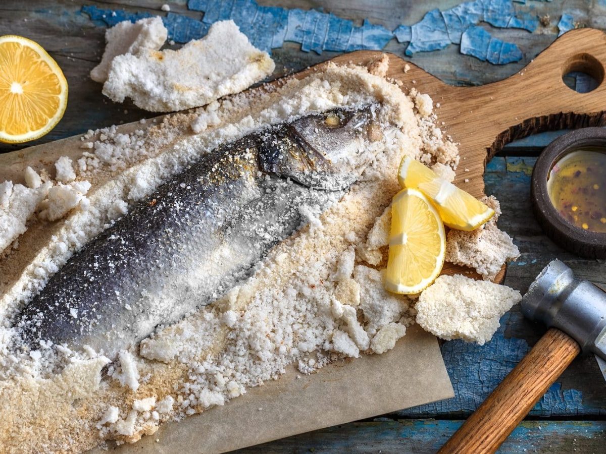 Aus der Vogelperspektive: Auf einem Holzbrett liegt ein Fisch in einer Salzkruste. Daben lieben ein Beil, Zitronenhälften, Salz und Pfeffer