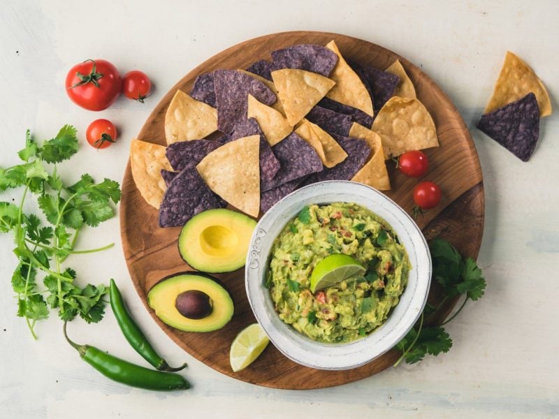 Guacamole auf rundem Holzbrett mit schwarzen und gelben Nachos, aufgeschnittener Avocado auf heller Steinplatte. Daneben Chile, Tomaten und Petersilie. Aufnahme aus der Vogelperspektive.