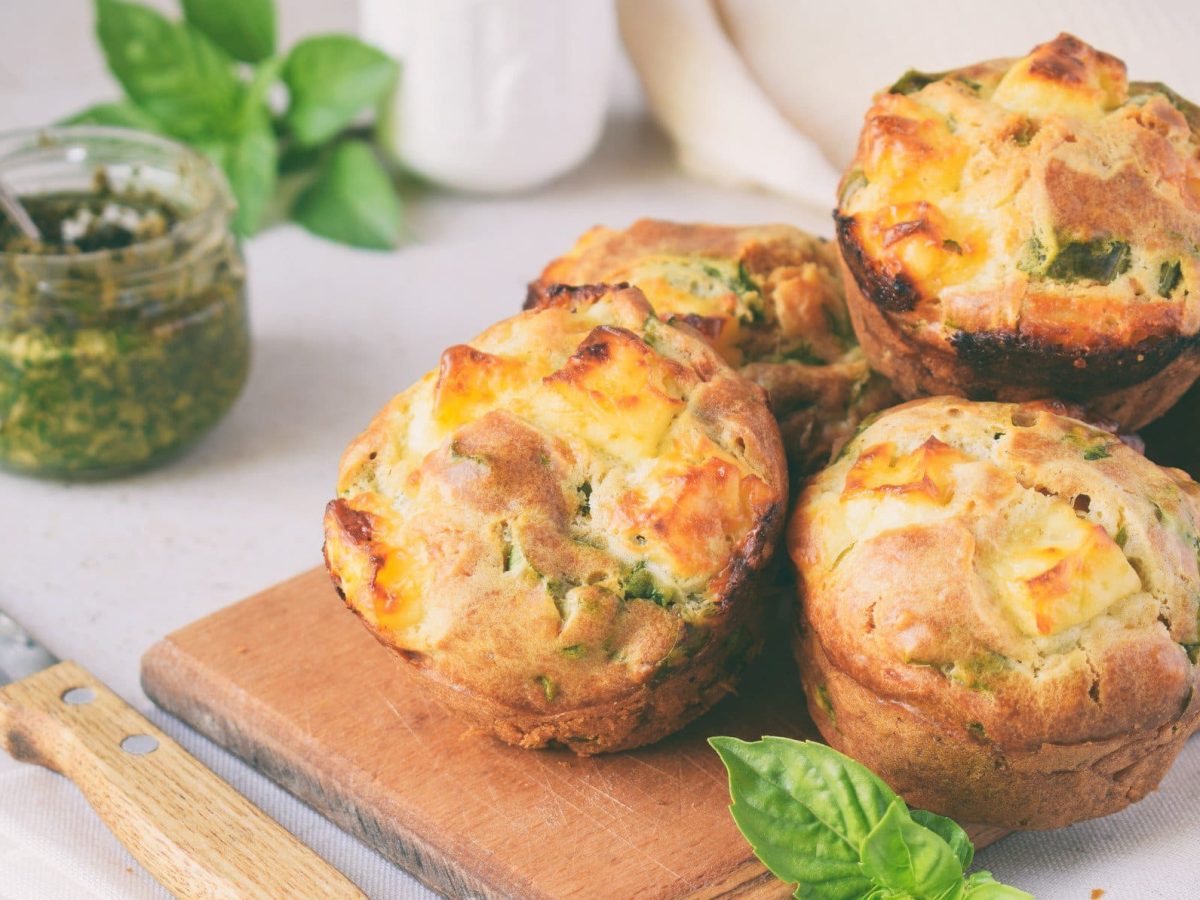 Glutenfreie Frühstücksmuffins auf einem Brett, daneben liegt ein Messer, außerdem ein Glas Pesto und einige Basilikumblätter.