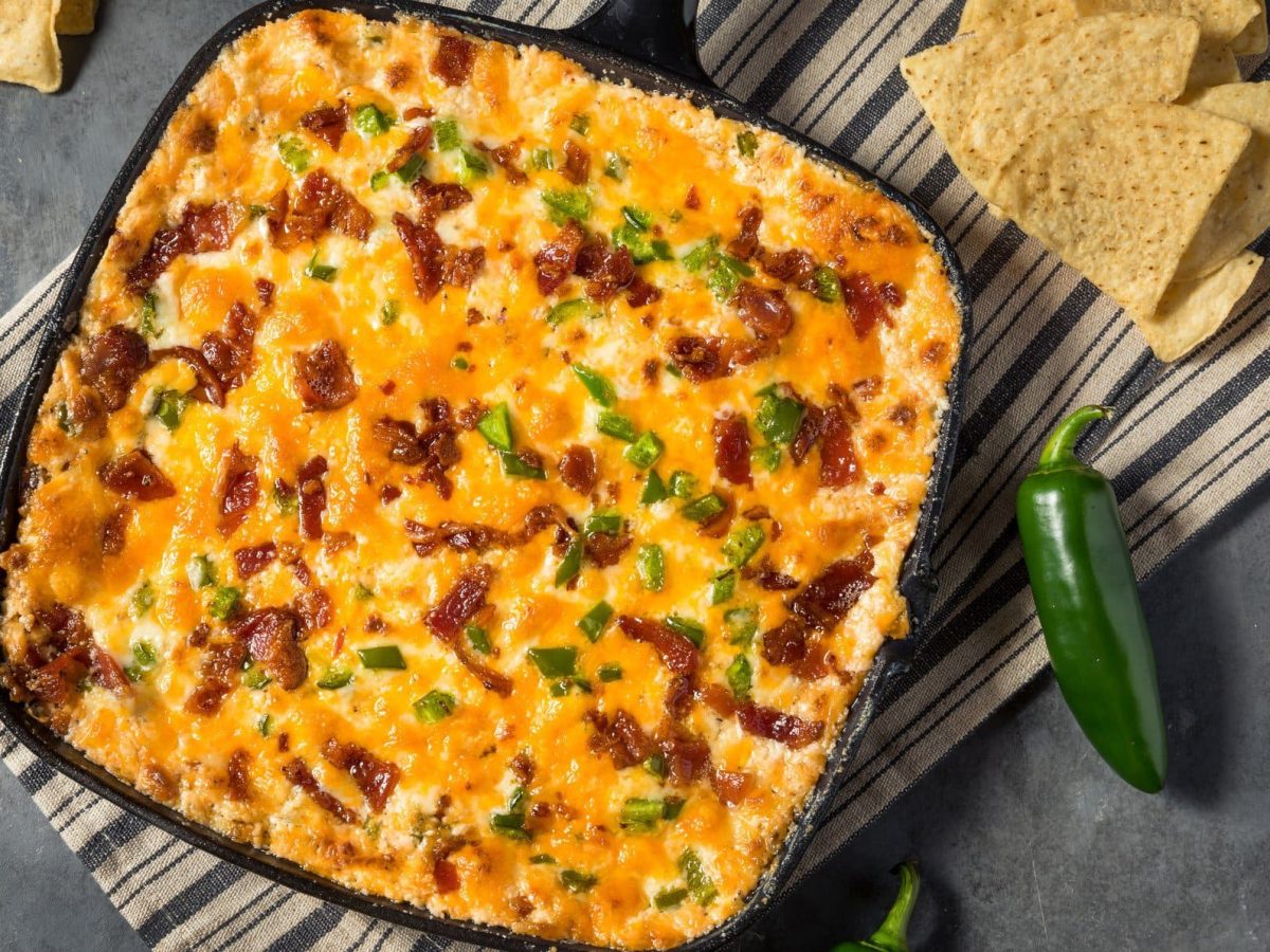 Jalapeno Popper Dip in Gusspfanne mit grüner Jalapeno und Nchos auf gestreifeter Stoffserviette auf grauem Untergrund. Daneben Nachos und Jalapenos. Aufnahme aus der Vogelperspektive.