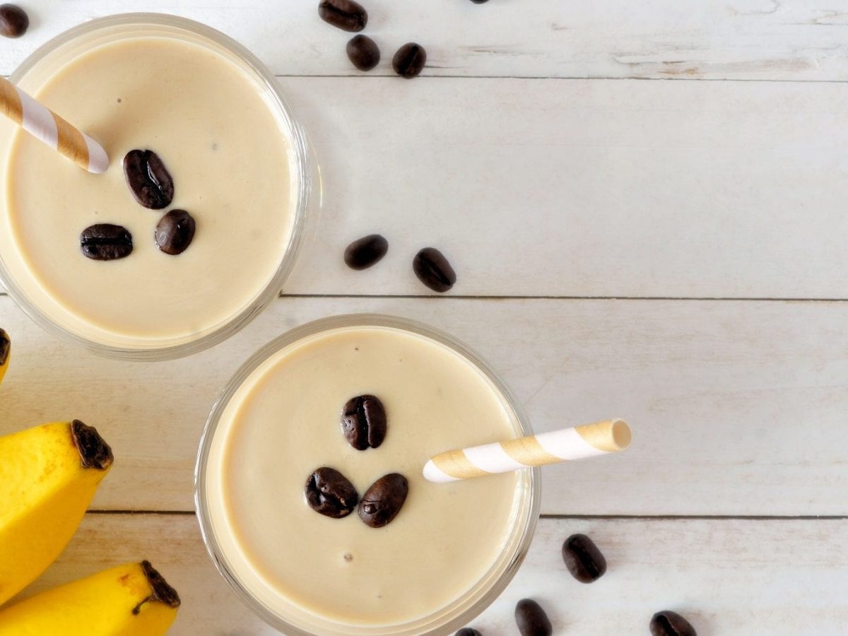 Zwei Gläser Kaffee-Bananen-Smoothie stehen auf einem weißen Holztisch. Kaffeebohnen und Bananen als Deko dazu.