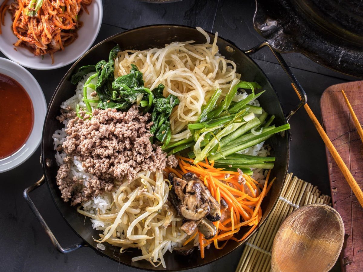 Koreanisches Bibimbap, bestehend aus Karotten, Pilzen, Rettich, veganem Hackfleisch, Gurke, Karotte und Sprossen auf einem Bett aus Reis in einer schwarzen Schale, dazu Chili-Sauce und Kimchi