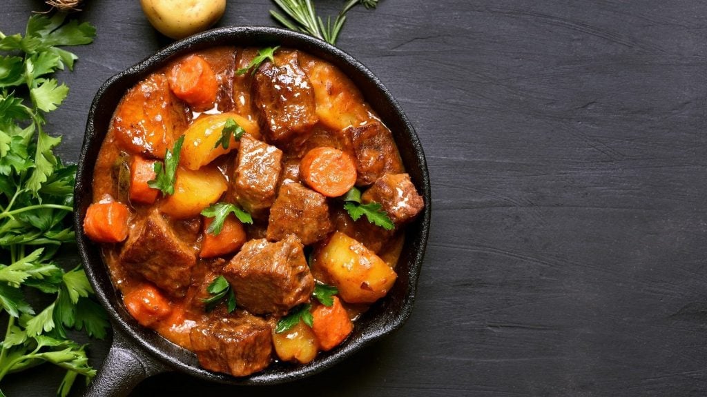 Aus der Vogelperspektive ist ein Topf zu sehen, in dem Lammgulasch mit frischen Kräutern ist Neben dem Topf liegen eine Zwiebel, eine Kartoffel und ein Zweig Rosmarin.