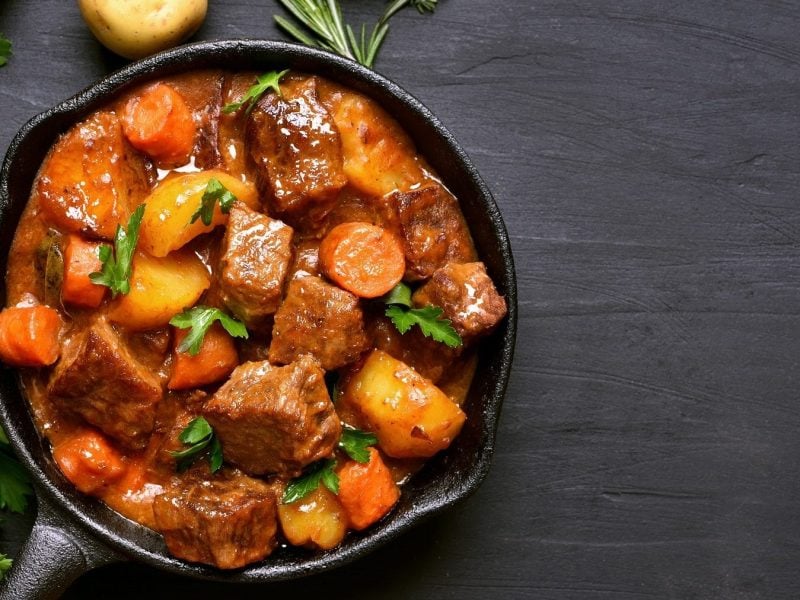 Aus der Vogelperspektive ist ein Topf zu sehen, in dem Lammgulasch mit frischen Kräutern ist Neben dem Topf liegen eine Zwiebel, eine Kartoffel und ein Zweig Rosmarin.