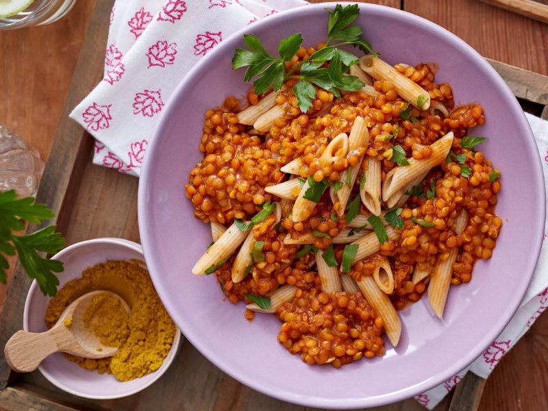 Linsen-Curry mit Pasta und frischer Petersilie in einer rosa Schale auf einem hölzernen Tisch