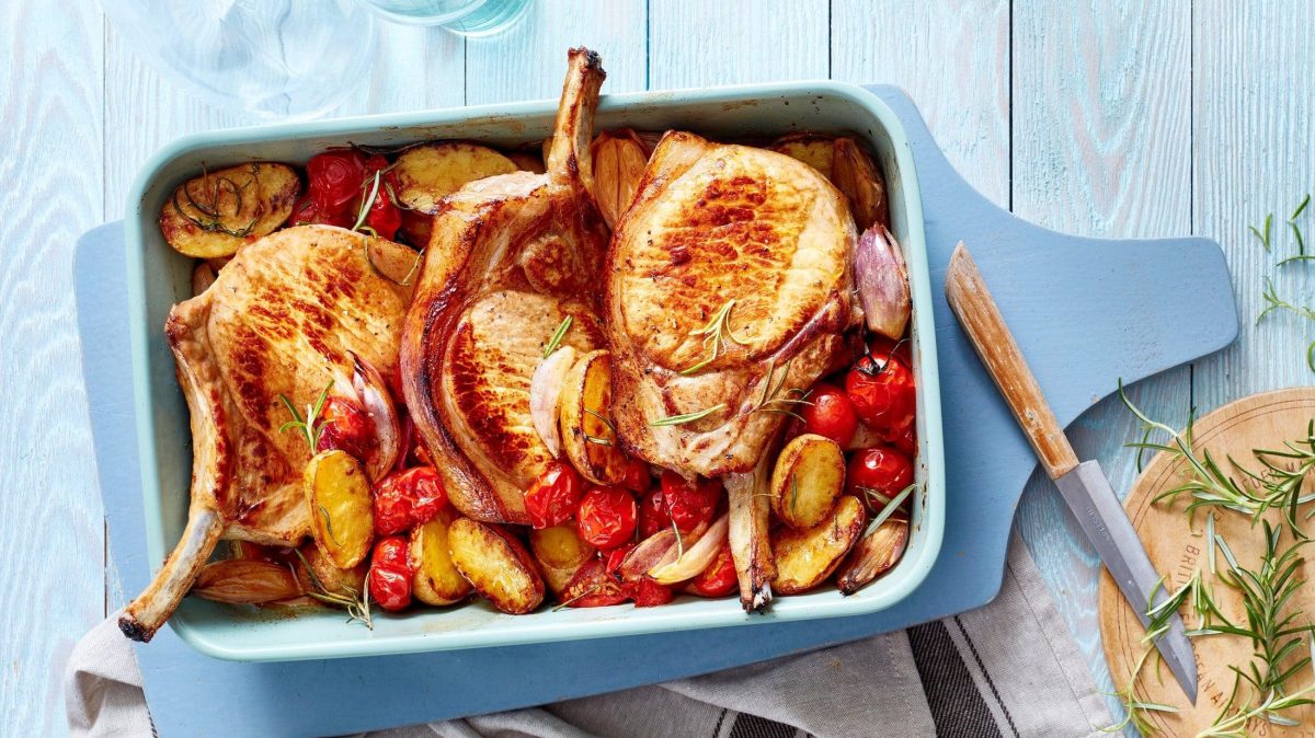 Die mediterranen Stielkoteletts mit Kartoffeln, Tomaten und Gewürzen stehen angerichtet auf dem Tisch in einer blauen Auflaufform.