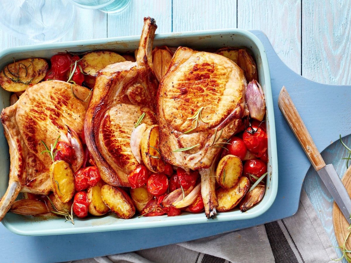 Die mediterranen Stielkoteletts mit Kartoffeln, Tomaten und Gewürzen stehen angerichtet auf dem Tisch in einer blauen Auflaufform.