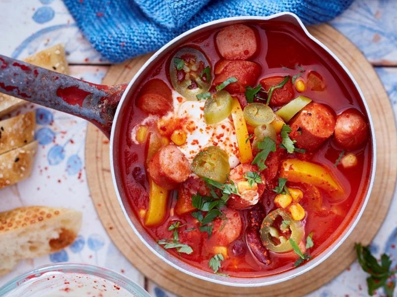 Ein mexikanisches Würstchen-Gulasch mit Paprika, Jalapeno, Tomaten, Würstchen und Crème Fraiche in einem Messingtopf auf einem Brettchen serviert