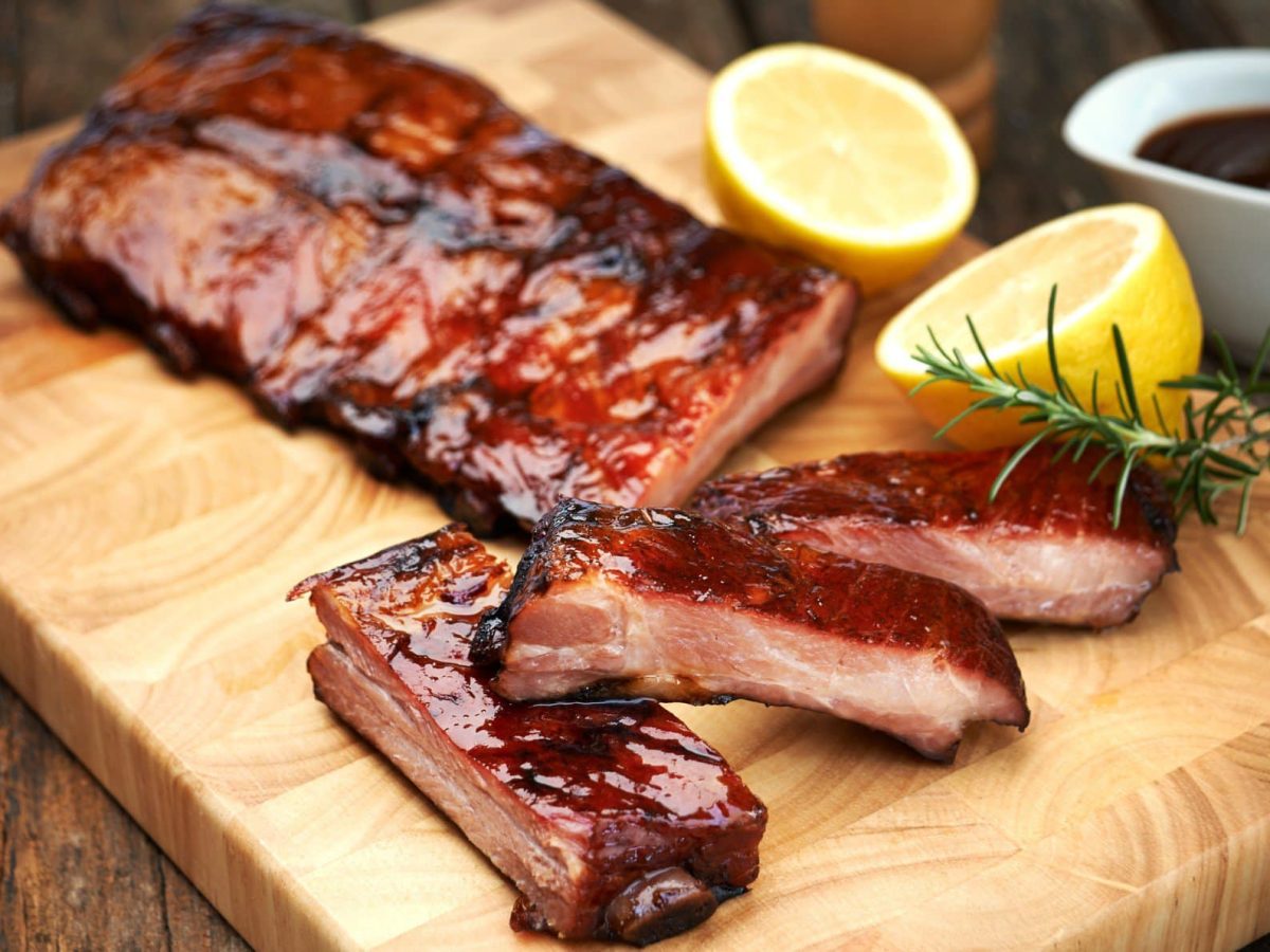 Ofen Spareribs auf Holzbrett mit Dillzweig und Zitronenhälften. Daneben Schale mit Sauce. Angerichtet auf dunklem Holztisch. Frontalaufnahme.