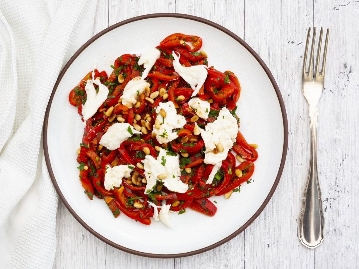 Aus der Vogelperspektive: Auf einem weißen Untergrund steht ein weißer Teller mit gerösteter Paprika mit Mozzarella und Pinienkernen. Daneben liegen ein weißes Tuch und eine Gabel.