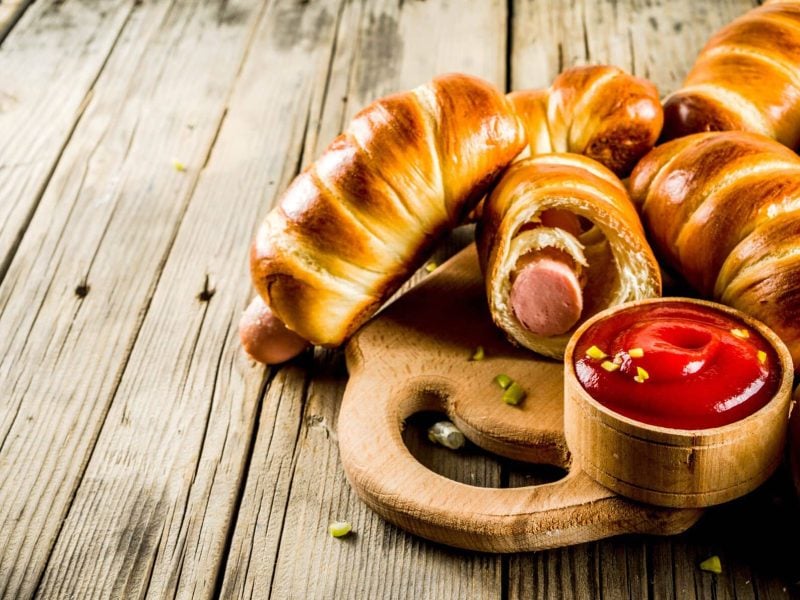 Pigs in Blankets zum Superbowl auf Holzbrett und Holztisch. Eines angeschnitten. Eine Schale mit Ketchup im Verdergrund und Würfel vo sauren Gurken als Deko verteilt. Frontalansicht.