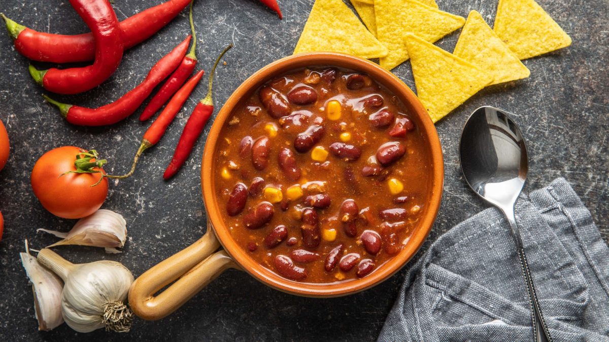 Ein Topf pikante Bohnensuppe mit Mais in der Draufsicht, daneben ein Löffel, Chilischoten, Knoblauch und Nachos.