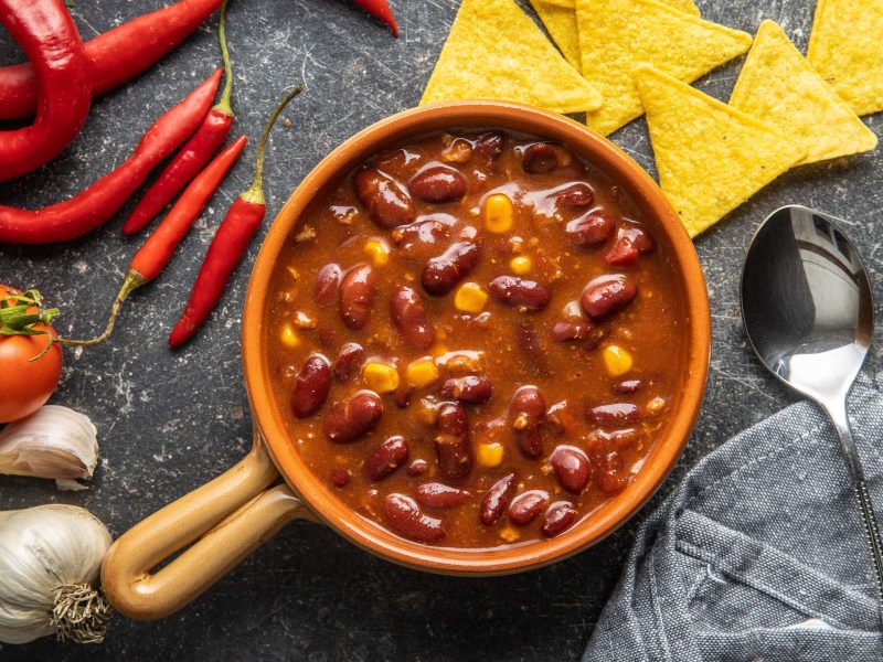 Ein Topf pikante Bohnensuppe mit Mais in der Draufsicht, daneben ein Löffel, Chilischoten, Knoblauch und Nachos.