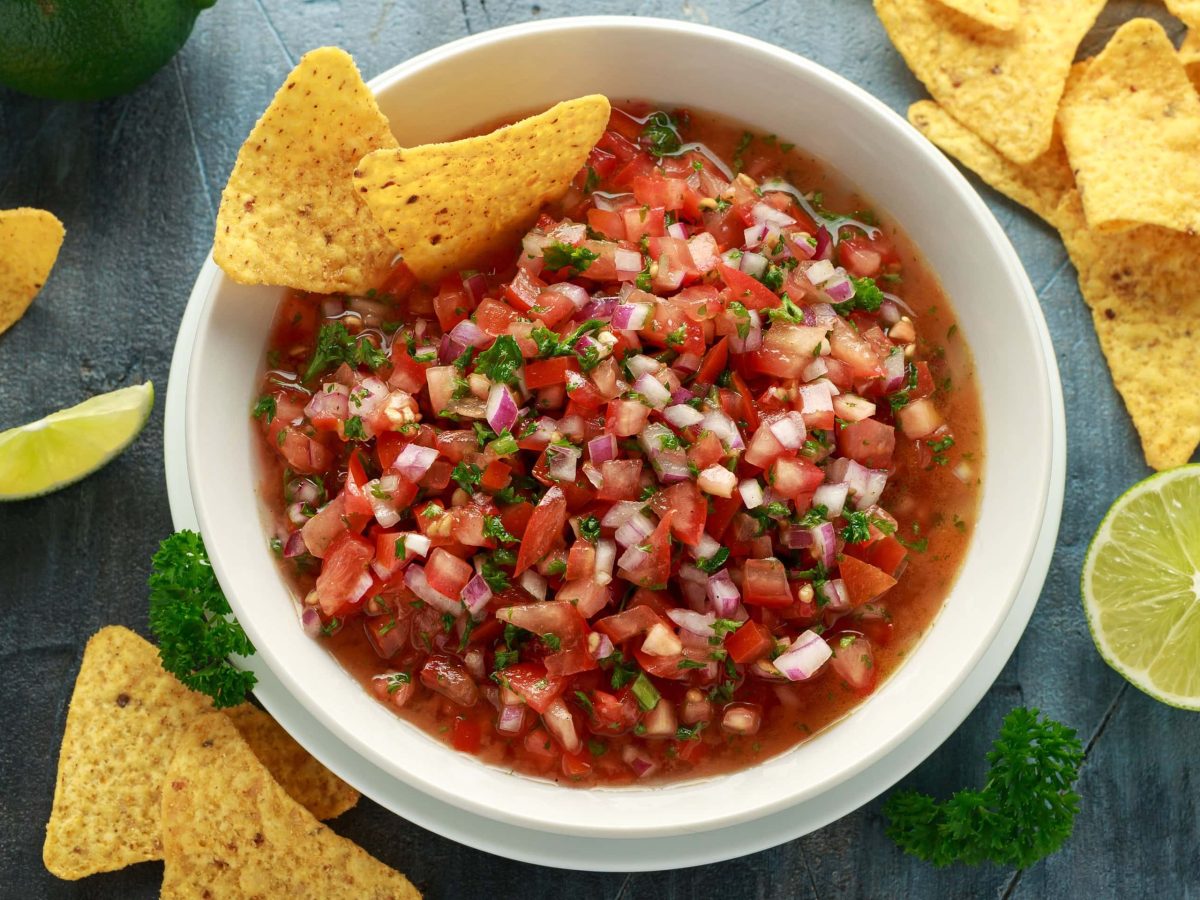 Eine Schüssel Salsa mit Nachos in der Draufsicht, daneben liegen weitere Chips und Limetten.