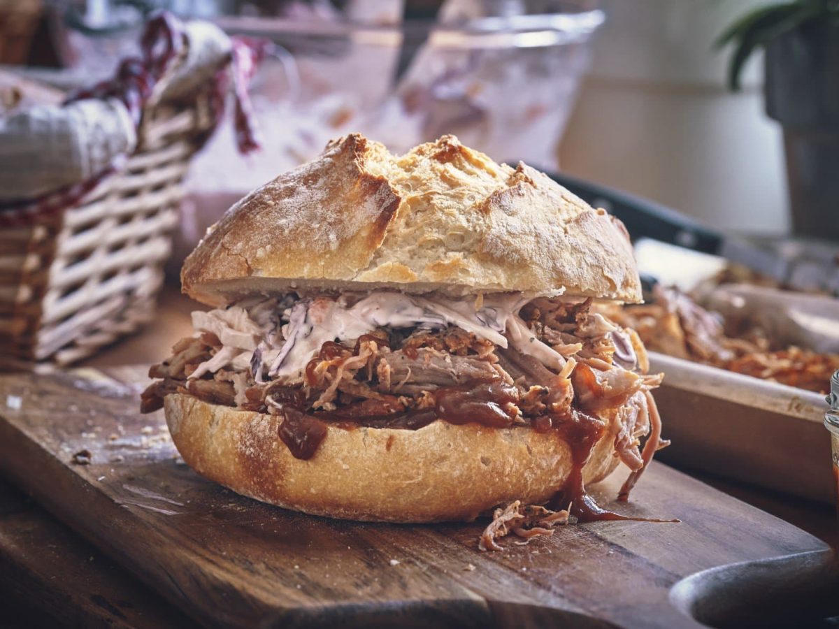 Ein Pulled-Pork-Sandwich mit Coleslaw auf einem Holzbrett. Im Hintergrund verschiedene Zutaten.