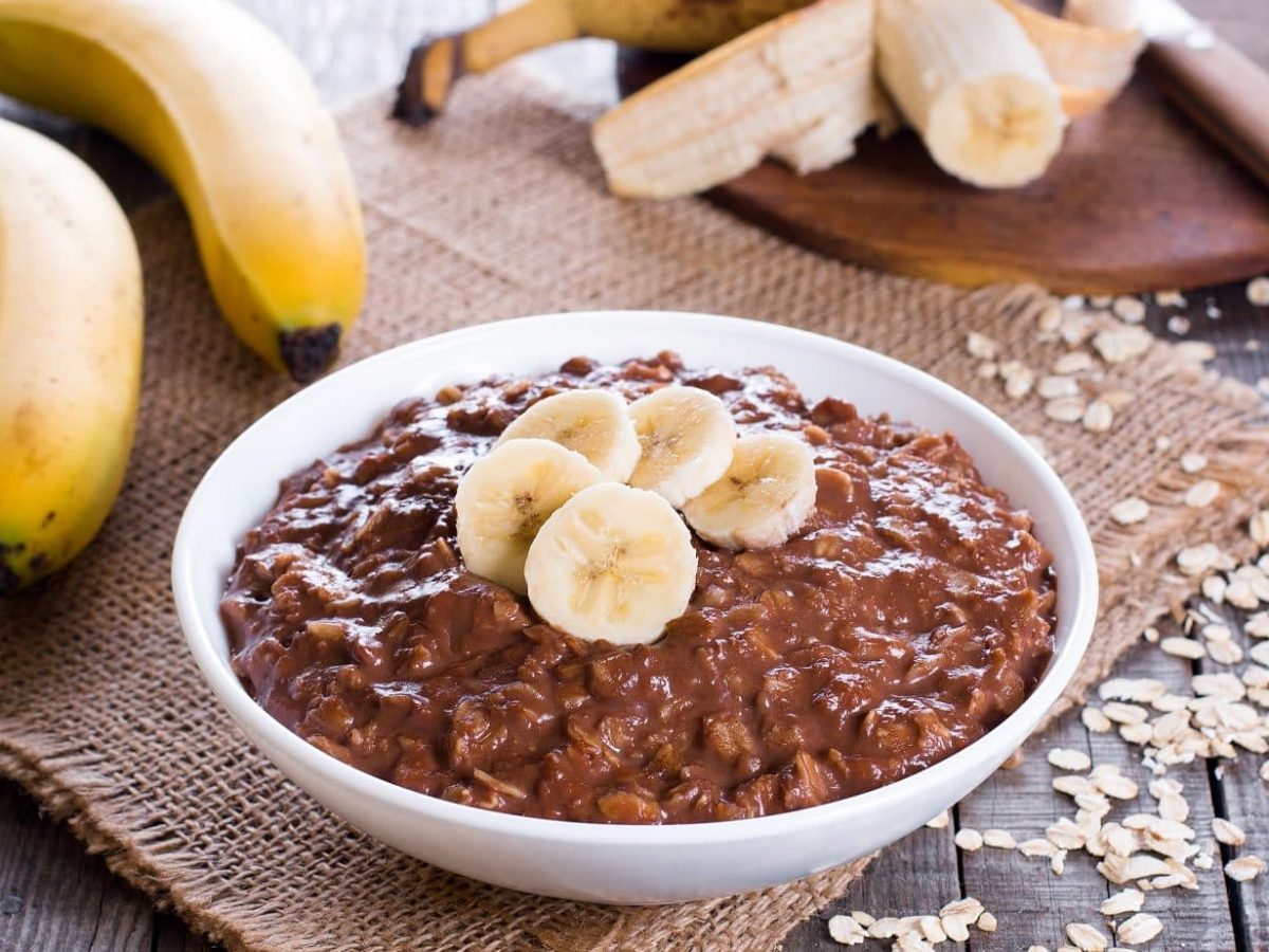 Eine Schale voll von unserem Schokoladen-Porridge steht auf dem Tisch. Im Hintergrund Bananen, Haferflocken als Deko,