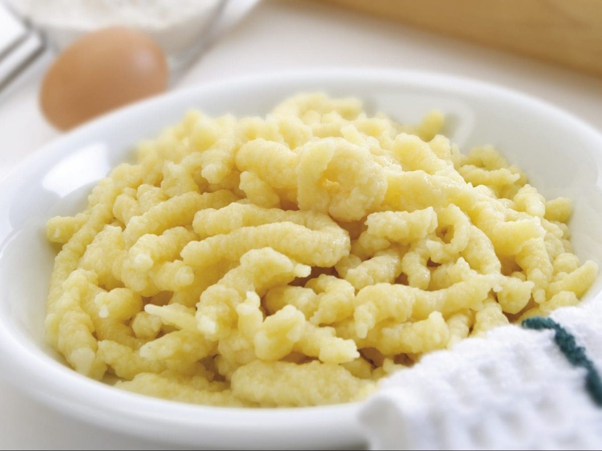 Ein weißer Teller mit selbstgemachten Spätzle steht auf einem Tisch. Daneben liegen ein Ei, eine Schale mit Mehl und ein Handtuch.