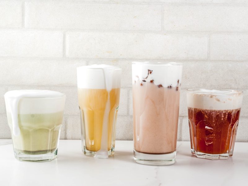 Vier Gläser mit verschiedenfarbigen Cheese-Tea-Sorten stehen auf einem weißen Tisch vor einer weißen Wand.