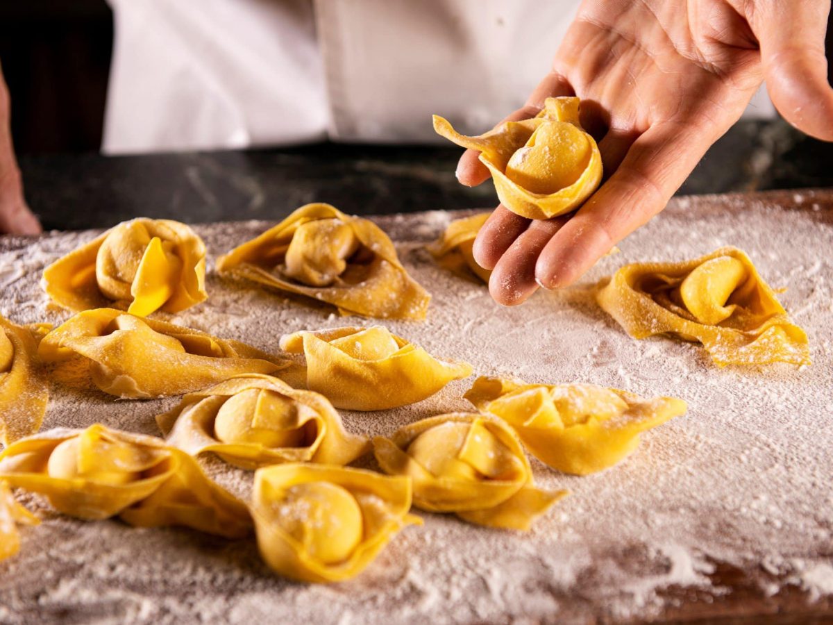 Handgemachte Tortellini auf mit Mehl bestäubtem Tisch. Hände und Kochjacke eines Koch im Hintergrund sichtbar. In der linken Hand hält er eine Tortellini. Frontalaufnahme zum Tag der Tortellini.