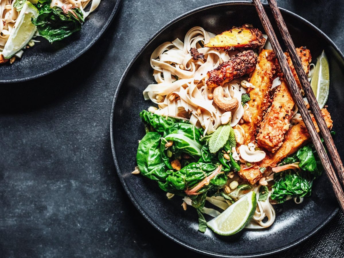 Tempeh-Spinat-Pfanne mit Reisbandnudeln, Kokosmilch, Limetten und gerösteten Nüssen in einer großen Schale auf Schieferuntergrund