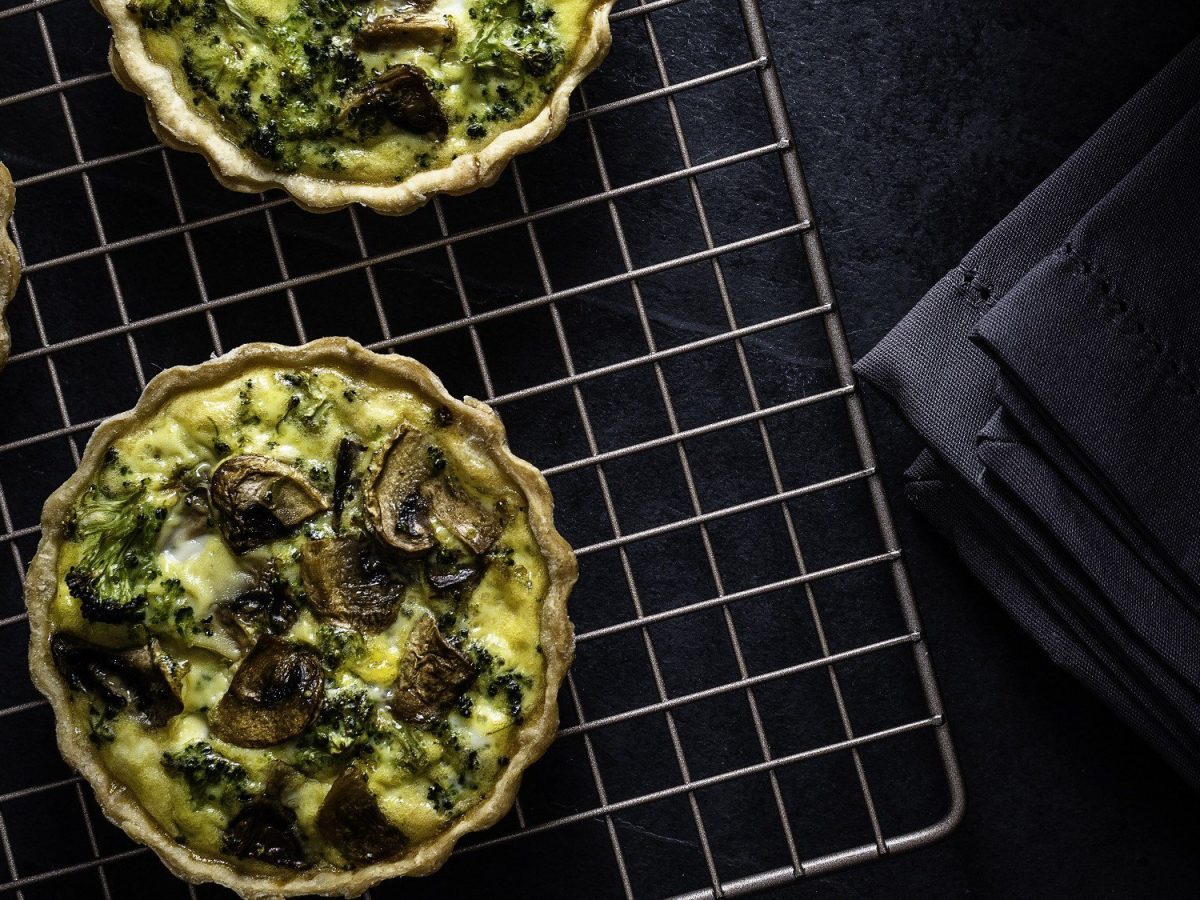 Saftige vegane Mini-Quiches mit cremiger Pilz-Brokkoli-Füllung auf einem Gitter vor schwarzem Hintergrund. Aufnahme aus der Vogelperspektive..