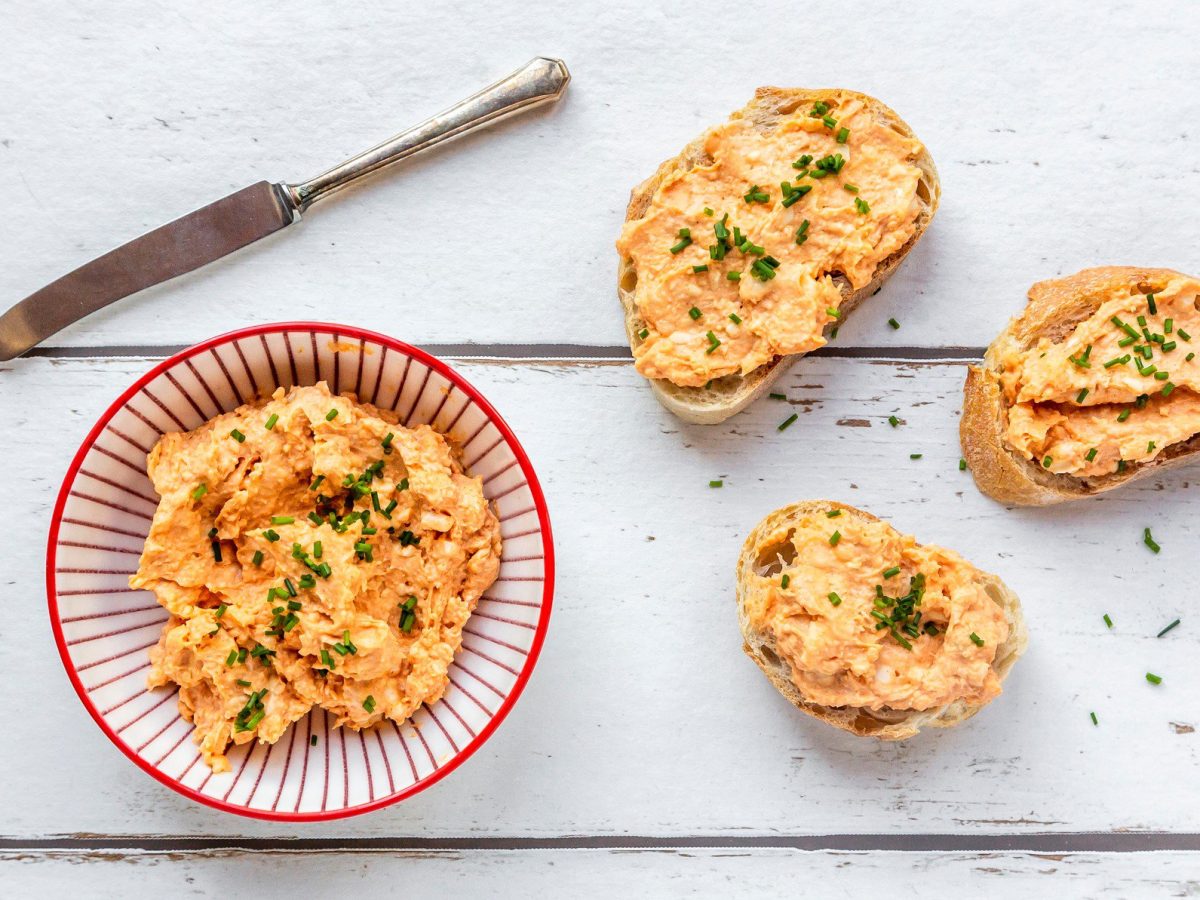 Eine Schüssel veganer Obazda mit einigen bestrichenen Baguettescheiben und einem Messer in der Draufsicht.