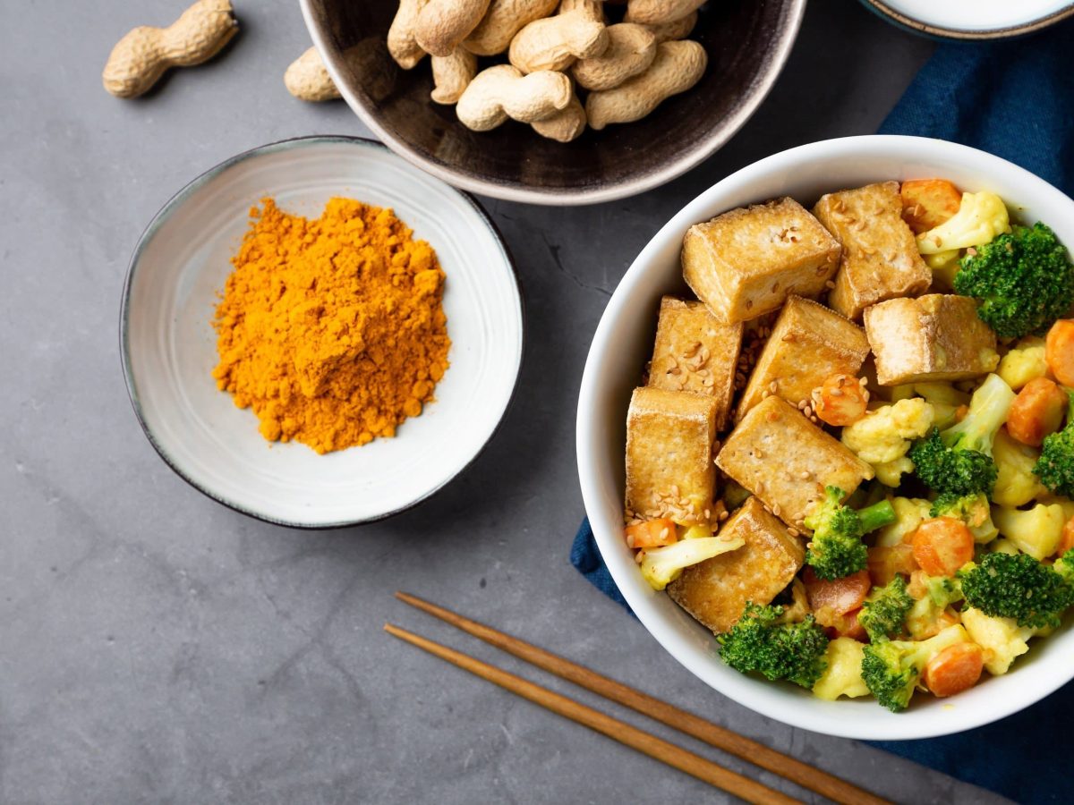 Wok-Gemüse in Erdnusssauce, bestehend aus Brokkoli, Blumenkohl, Karotten, serviert mit Tofuwürfeln in einer weißen Bowl auf dunklem Untergrund