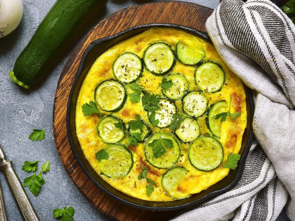 Aus der Vogelperspektive: Eine runde Zucchini-Frittata. Es handelt sich um eine Eierspeise. Neben der runden Form liegen ein handtuch, zwei Gabeln und frische Kräuter