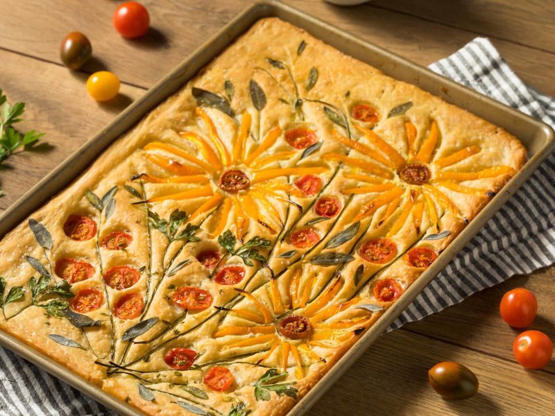 Flower-Focaccia mit floralen Applikationen aus Kräutern und Tomaten auf einem Holztisch.