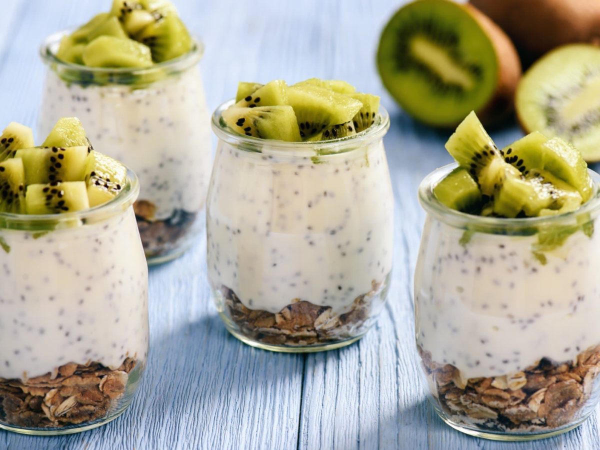 Drei Gläser Chia-Joghurt mit Müsli und Kiwi auf hellblauem Untergrund aus Holz.