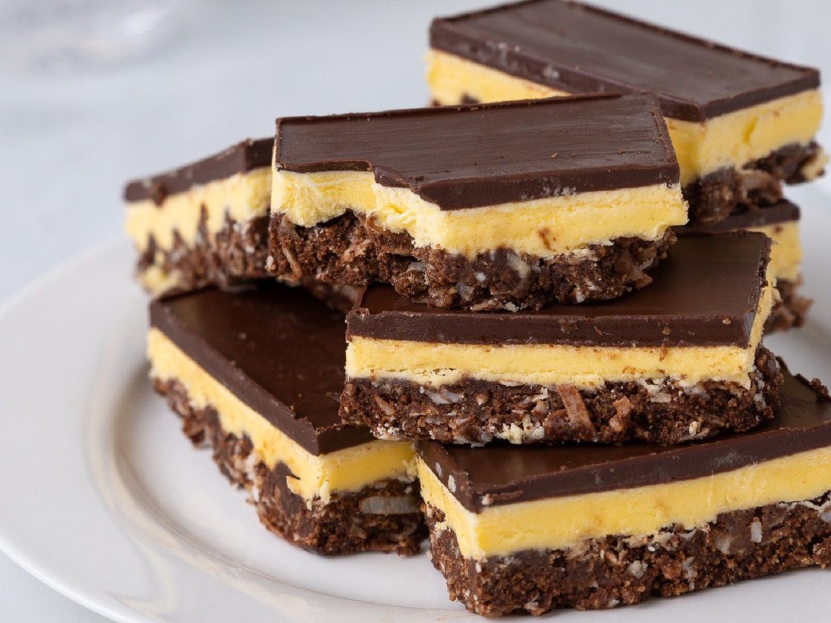 Auf einem Teller liegen mehrere Stücken selbstgemachter Nanaimo Bars. Sie bestehen aus einem Keksboden, einer Creme und einer Schokoschicht.