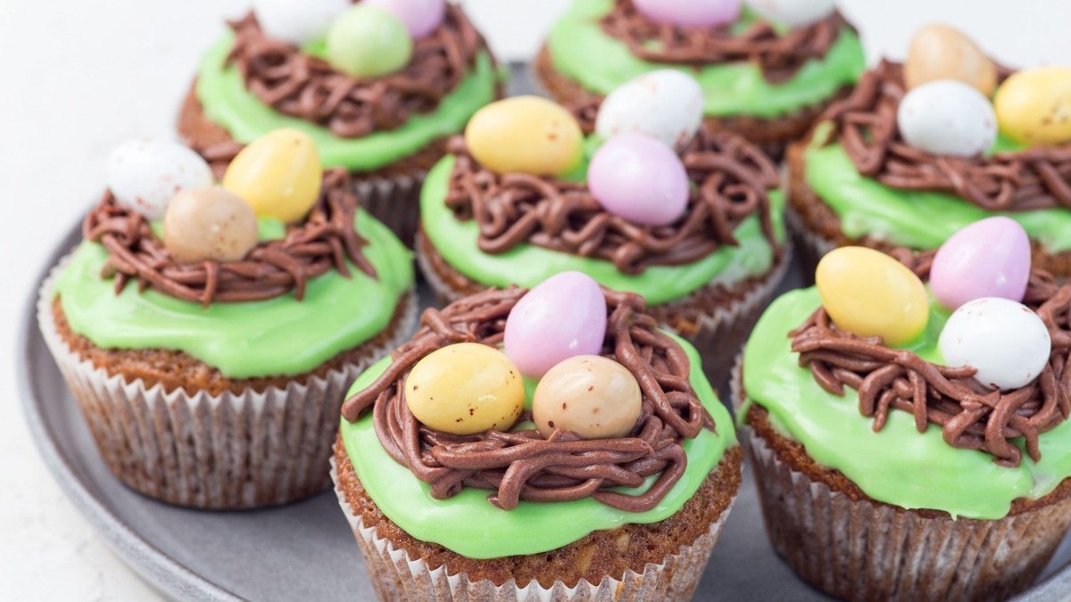 Auf einem Teller liegen mehrere Oster Muffins. Sie sind mit Zuckereiern und ein einem grünen Frosting dekoriert. Die Dekoration sieht aus wie Osterei-Nester.