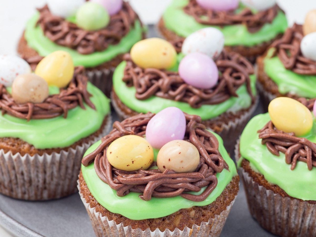 Auf einem Teller liegen mehrere Oster Muffins. Sie sind mit Zuckereiern und ein einem grünen Frosting dekoriert. Die Dekoration sieht aus wie Osterei-Nester.