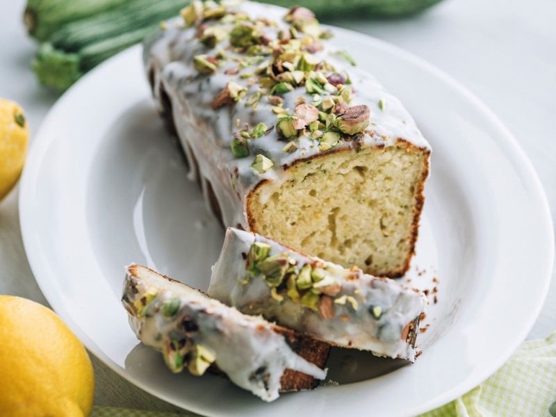 Auf einem Teller liegt ein Zucchini-Kuchen mit Zitrone aus der Kastenform. Er ist übezogen mit einer Zuckerglasur und Pistazien.