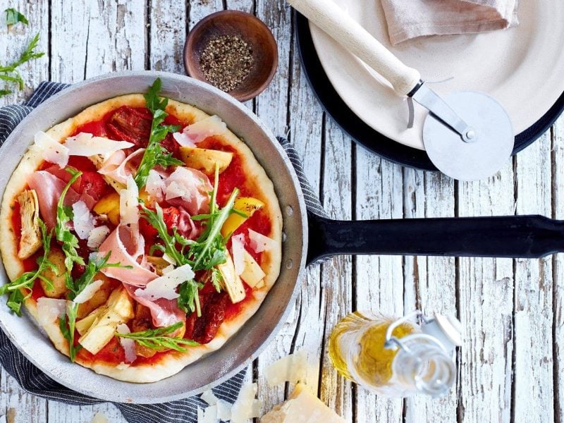 In einer Pfanne liegt eine fertige Pizza mit Antipasti. Sie ist verfeinert mit Schinken, Rucola und Parmesan.