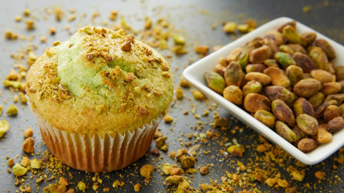 Ein Pistazien-Muffin steht auf einer Schiefertafel. Daneben liegen Pistazien in einer Schale.