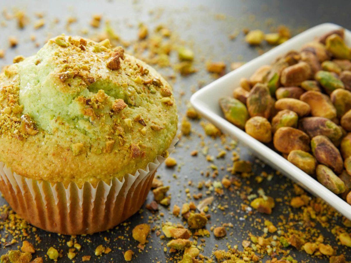 Ein Pistazien-Muffin steht auf einer Schiefertafel. Daneben liegen Pistazien in einer Schale.