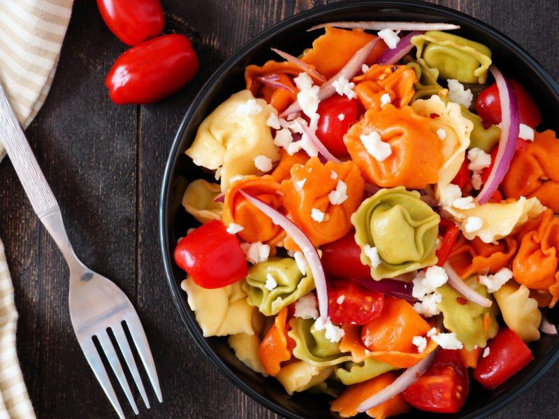 Unser bunter Tortellini-Salat steht angerichtet in einer Bowl auf einem braunen Holztisch.