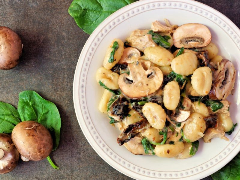 Gnocchi-Pfanne mit Pilzen und Spinat auf weißem Teller, angerichtet auf grauem Untergrund. Umgeben ist der Teller von Champignons und Spinat.