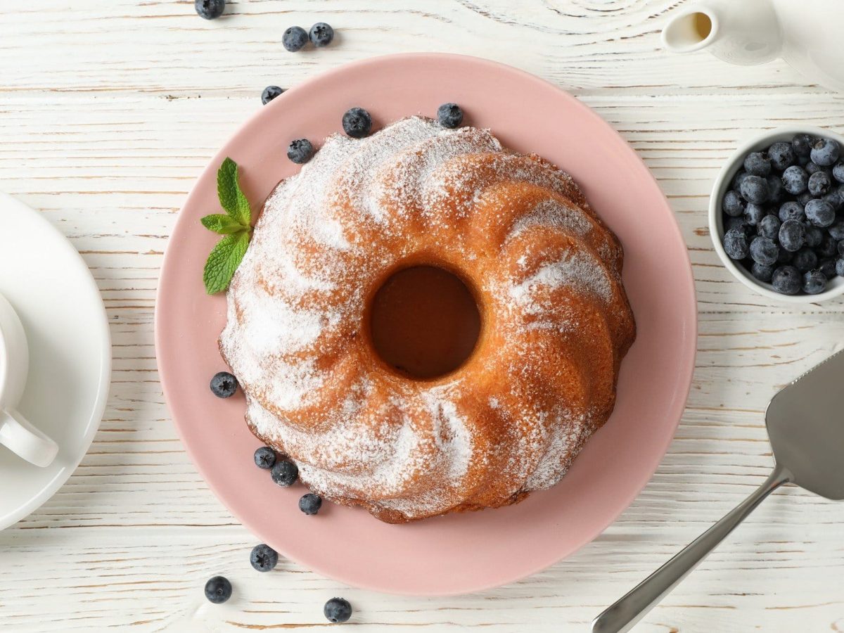 Gugelhupf mit Puderzucker auf rosa Teller. Dieser steht auf einem weißen Holztisch, umgeben von Heidelbeeren und einer Tasse Tee mit Zitrone.