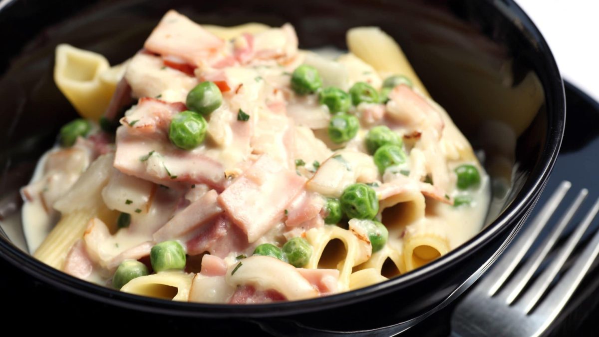 Penne Alfredo mit Kochschinken und Erbsen in schwarzer Schüssel. Daneben liegt eine Gabel auf einem schwarzen Unterteiller.