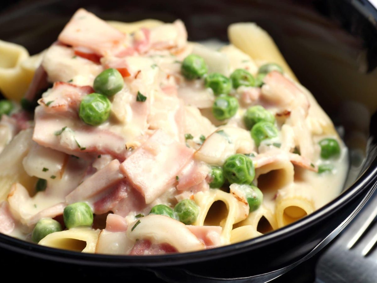 Penne Alfredo mit Kochschinken und Erbsen in schwarzer Schüssel. Daneben liegt eine Gabel auf einem schwarzen Unterteiller.